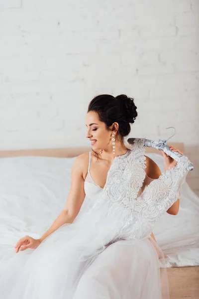 Noiva em sutiã e brincos de pérola segurando cabide com vestido de noiva branco na cama em casa — Fotografia de Stock