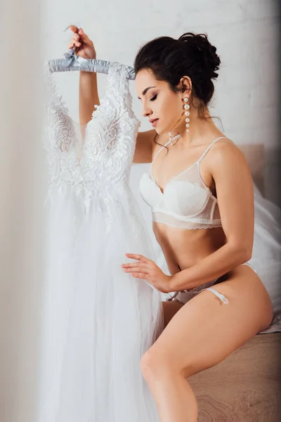 Selektiver Fokus der brünetten Braut in Unterwäsche berührt weißes Hochzeitskleid auf Kleiderbügel im Schlafzimmer — Stockfoto