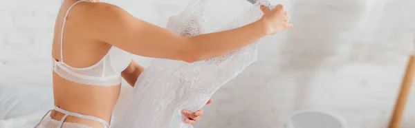 Panoramic shot of bride in lingerie holding white lace wedding dress on hanger — Stock Photo