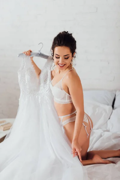 Sexy bride in underwear and garter belt touching wedding dress on hanger on bed — Stock Photo