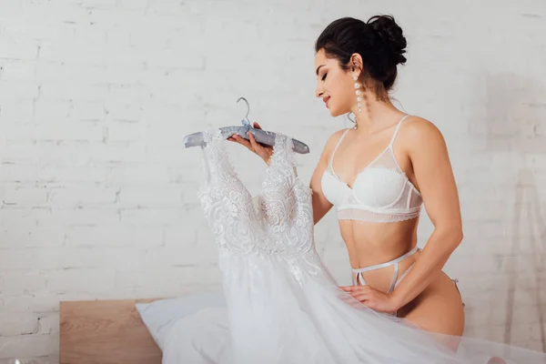 Seductive bride in lingerie and pearl earrings holding hanger with lace wedding dress on bed — Stock Photo