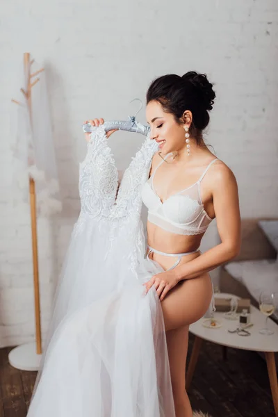 Брюнетка наречена в мереживному бюстгальтері тримає білу весільну сукню на вішалці вдома — стокове фото