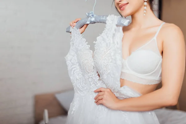 Cropped view of bride in bra holding sating hanger with lace wedding dress at home — Stock Photo