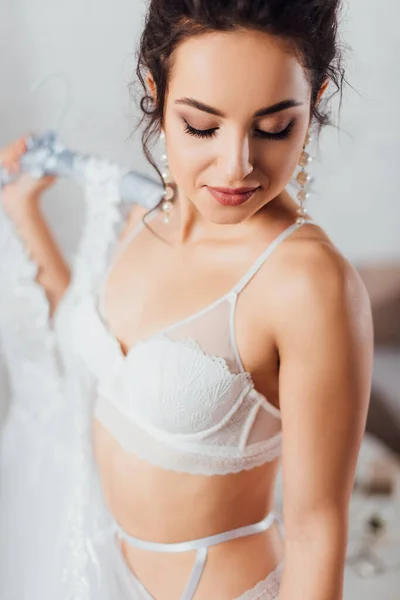 Selective focus of bride in bra and pearl earrings holding hanger with wedding dress — Stock Photo