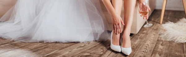 Panorâmica tiro de noiva em sapatos de casamento segurando copo de vinho perto do vestido em casa — Fotografia de Stock