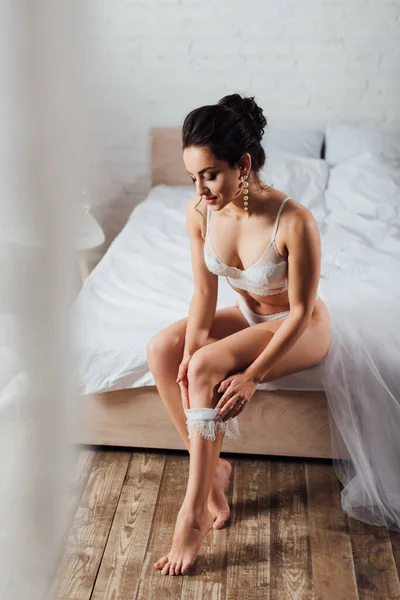 Selective focus of bride in white lingerie wearing wedding garter while sitting on bed — Stock Photo