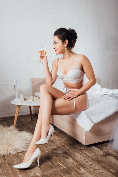 Sposa in reggiseno e scarpe che reggono un bicchiere di vino e distolgono lo sguardo sul letto — Foto stock