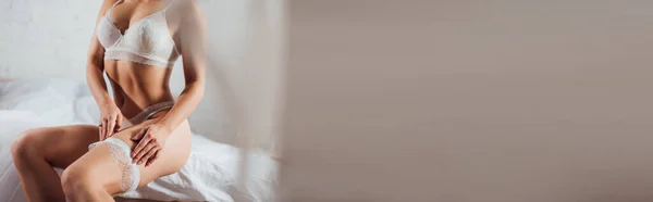 Panoramic shot of sexy bride putting on wedding garter in bedroom — Stock Photo