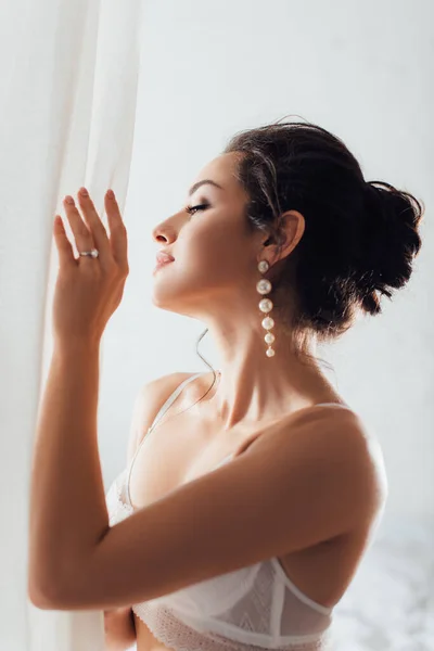 Selective focus of brunette bride in bra and pearl earrings touching white curtain at home — Stock Photo