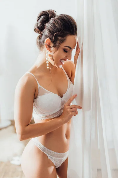 Sposa in lingerie di pizzo e orecchino di perle in piedi vicino alla tenda a casa — Foto stock