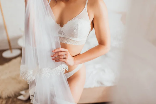Vue recadrée de la mariée en sous-vêtements blancs tenant voile de dentelle dans la chambre — Photo de stock