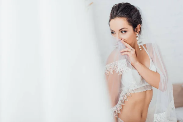 Selective focus of brunette bride in bra holding lace veil while looking away at home — Stock Photo