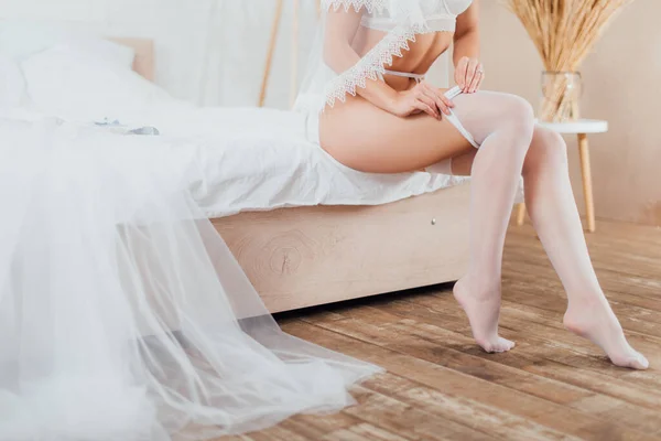 Cropped view of bride in bra and veil wearing stockings near wedding dress on bed — Stock Photo