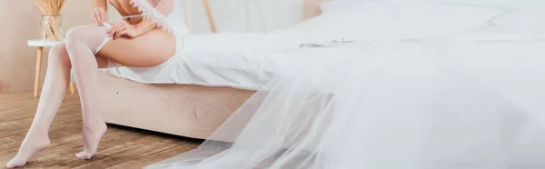 Panoramic crop of bride in veil and garter belt wearing stockings near wedding dress on bed — Stock Photo