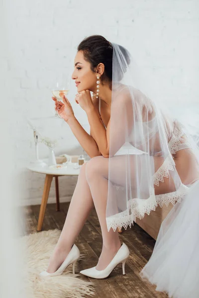 Focus selettivo della sposa in biancheria intima e velo in possesso di un bicchiere di vino vicino abito da sposa sul letto — Foto stock