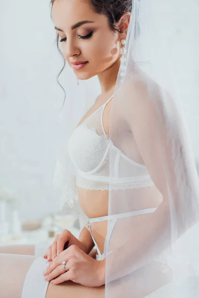 Noiva jovem em véu, roupa interior e cinto de liga vestindo meia branca em casa — Fotografia de Stock