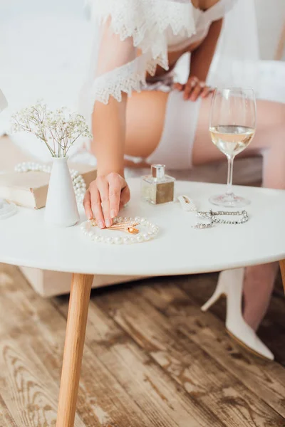 Selektiver Fokus der Braut in BH und Schleier, die Accessoires in der Nähe von Wein und Parfüm auf dem Couchtisch berührt — Stockfoto