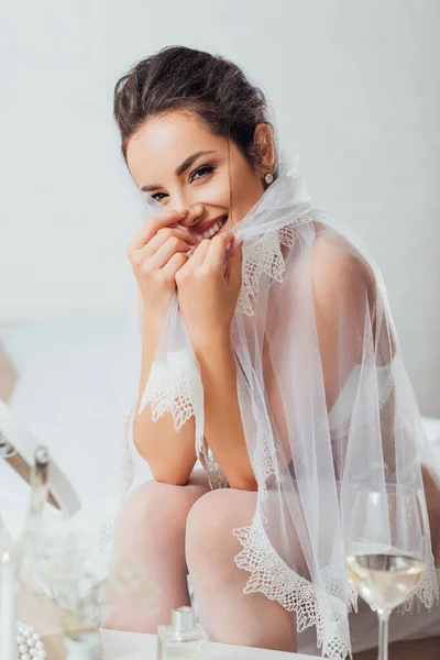 Selective focus of young bride holding veil while looking at camera near perfume, glass of wine and mirror — Stock Photo