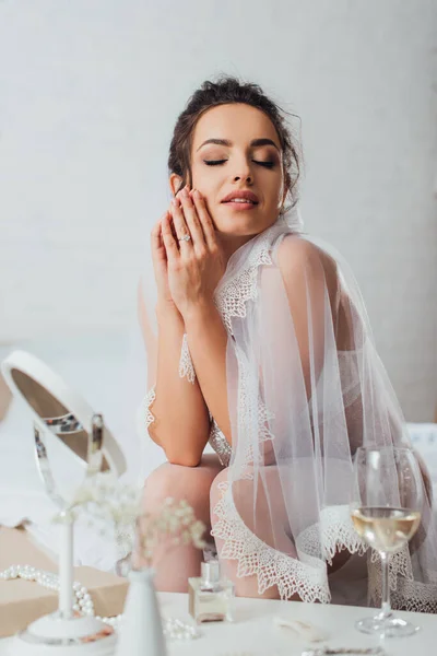 Focus selettivo della giovane sposa in reggiseno e velo seduta sul letto vicino al bicchiere di vino, specchio e profumo sul tavolino — Foto stock