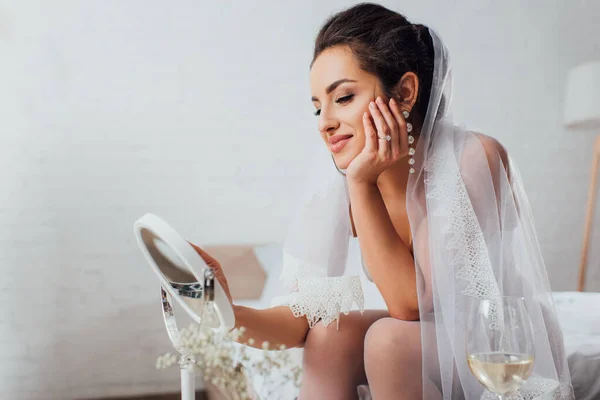 Selektiver Fokus der Braut in Schleier, die in den Spiegel neben einem Glas Wein im Schlafzimmer blickt — Stockfoto