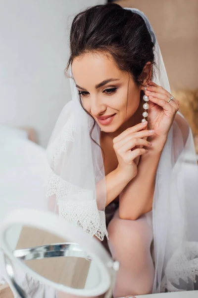 Selektiver Fokus der Braut in Schleier, die in die Kamera schaut, während sie zu Hause Perlenohrringe berührt — Stockfoto