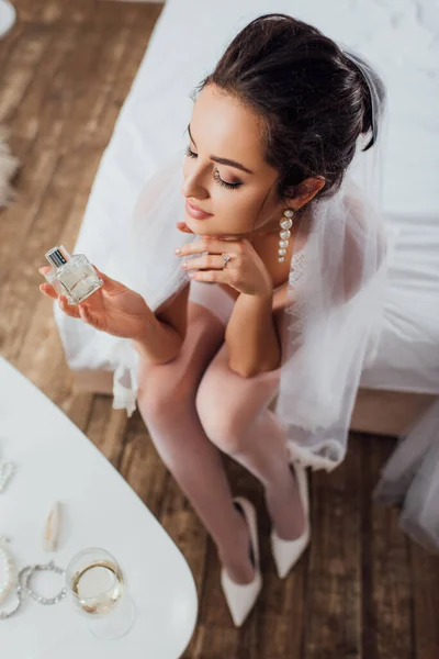 Вид сверху на невесту в вуали, держащую бутылку духов, сидя на кровати — стоковое фото