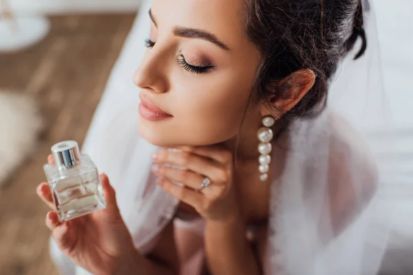 Foco seletivo da jovem noiva no véu segurando garrafa de perfume em casa — Fotografia de Stock
