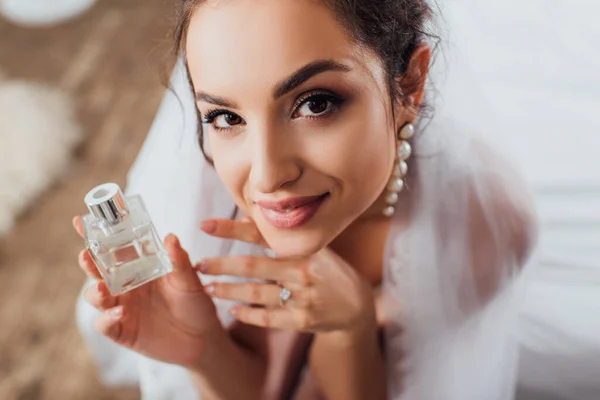 Vue grand angle de la jeune mariée voilée regardant la caméra tout en tenant le flacon de parfum sur le lit — Photo de stock