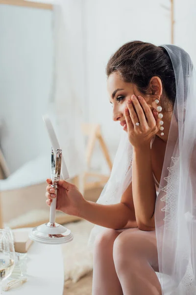 Concentration sélective de la mariée dans boucle d'oreille et voile tenant miroir près de la table basse à la maison — Photo de stock