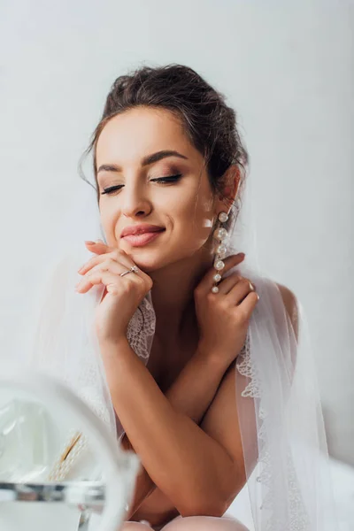 Selective focus of young bride with closed eyes touching lace veil near mirror — Stock Photo