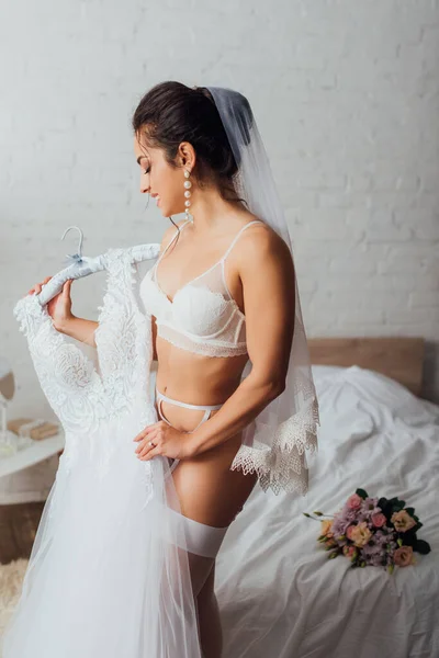 Сексуальная невеста в нижнем белье и вуали держа вешалку со свадебным платьем в спальне — стоковое фото