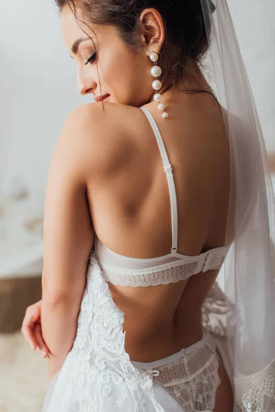 Focus selettivo della sposa in lingerie e velo che indossa abito da sposa bianco a casa — Foto stock