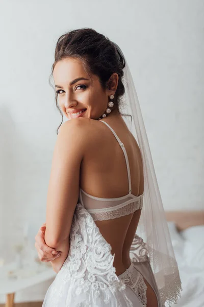 Selective focus of brunette bride in underwear, wedding dress and veil looking at camera — Stock Photo