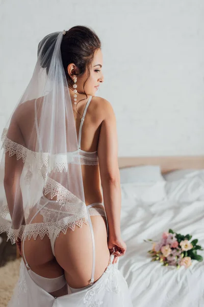 Vista trasera de novia sexy poniéndose vestido de novia blanco en el dormitorio - foto de stock