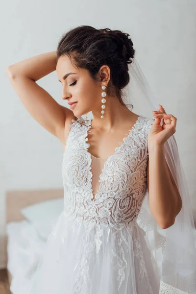 Jeune mariée avec les yeux fermés en dentelle robe de mariée touchant voile à la maison — Photo de stock