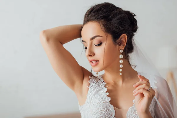 Selective focus of brunette bride in lace wedding dress touching veil — Stock Photo