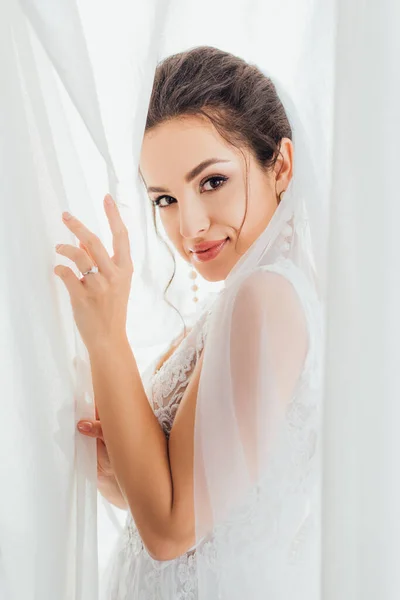 Vue latérale de la jeune mariée brune regardant la caméra tout en touchant les rideaux à la maison — Photo de stock