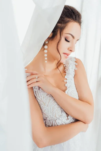 Foco seletivo da noiva em vestido de renda e brincos de pérola tocando ombro perto de cortinas — Fotografia de Stock
