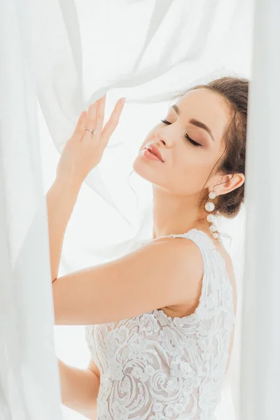 Selective focus of young bride with closed eyes touching white curtains — Stock Photo