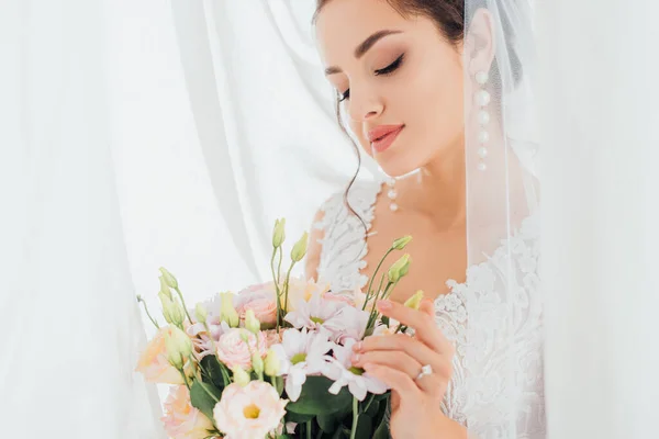 Foco seletivo da noiva tocando buquê perto de cortinas brancas — Fotografia de Stock