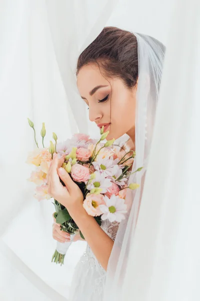 Selective focus of bride in veil holding flowers near white curtains — Stock Photo