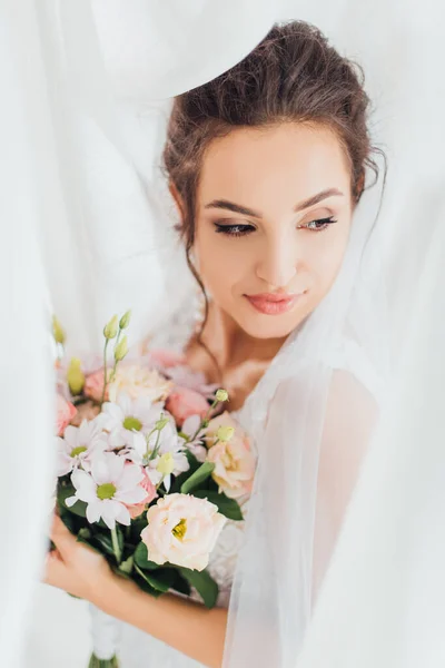 Выборочный фокус невесты брюнетки, смотрящей в сторону, держа цветочный букет возле белых занавесок — стоковое фото
