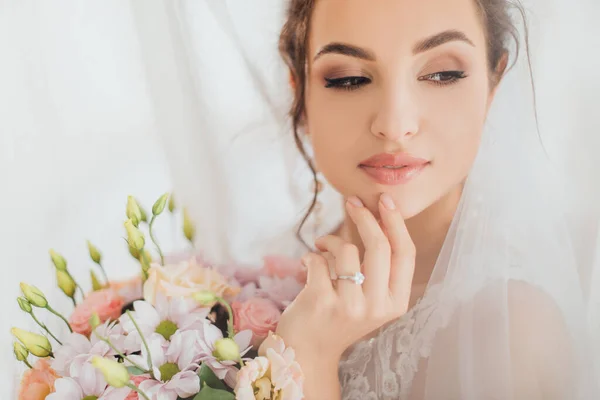 Concentration sélective de la mariée en voile toucher le menton tout en tenant bouquet près des rideaux — Photo de stock