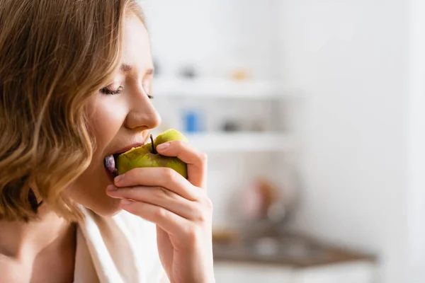 Jeune femme aux yeux fermés mordant pomme dans la cuisine — Photo de stock