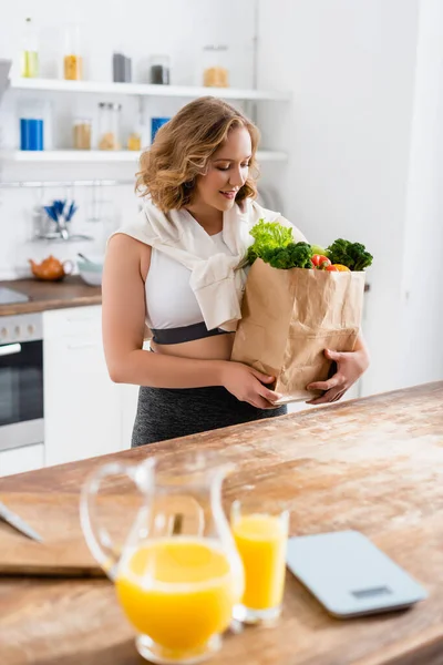 Selective focus of woman looking at paper bag with groceries near jug and glass with orange juice — Stock Photo