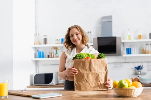 Young woman holding paper bag with vegetables near fruits in bowl — Stock Photo
