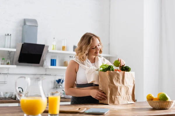 Enfoque selectivo de la mujer joven sosteniendo manzana cerca de la bolsa de papel con verduras y frutas en un tazón - foto de stock