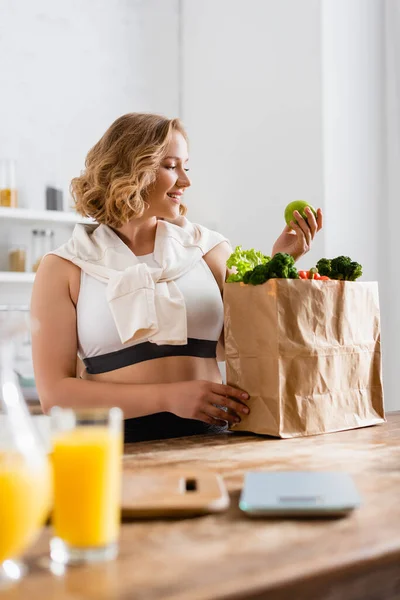 Fuoco selettivo della donna che tiene mela vicino al sacchetto di carta con verdure e brocca con succo d'arancia — Foto stock