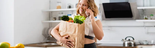 Récolte panoramique de jeune femme tenant sac en papier avec des légumes et parler sur smartphone — Photo de stock