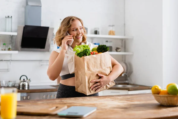 Foco seletivo da mulher segurando saco de papel com legumes e falando no smartphone — Fotografia de Stock